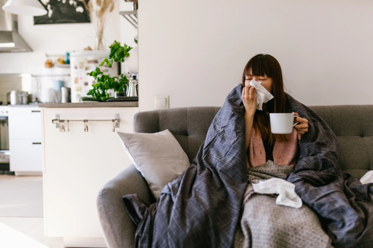 A sick employee sitting on a couch at home, wrapped in a blanket, holding a mug, and using tissues, representing the importance of paid sick leave.