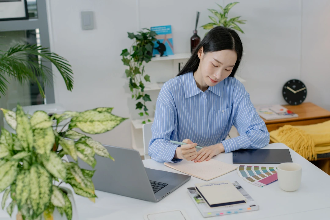 Woman working in a calm, plant-filled workspace while journaling—an example of a peaceful environment and daily habits that help relieve work stress.