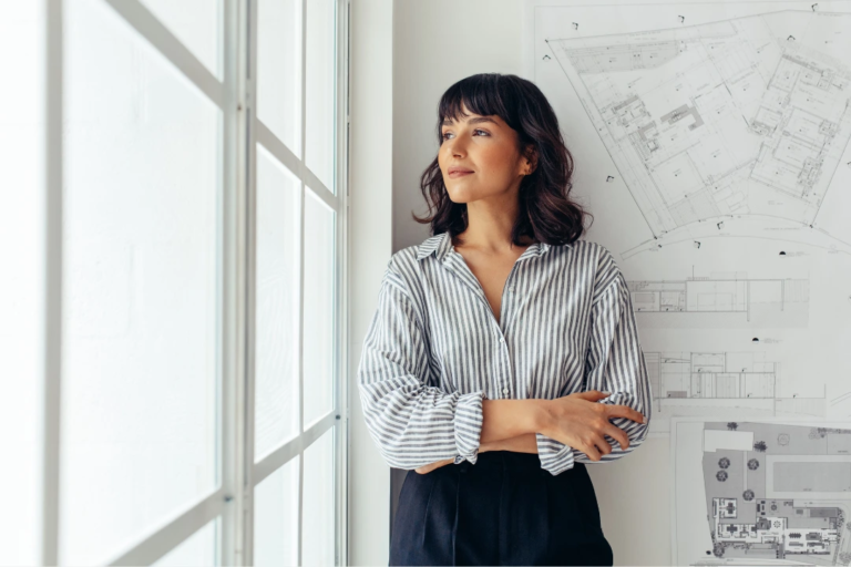 Professional woman standing peacefully by a window with architectural plans behind her, reflecting a moment of mindfulness and focus during a busy workday.