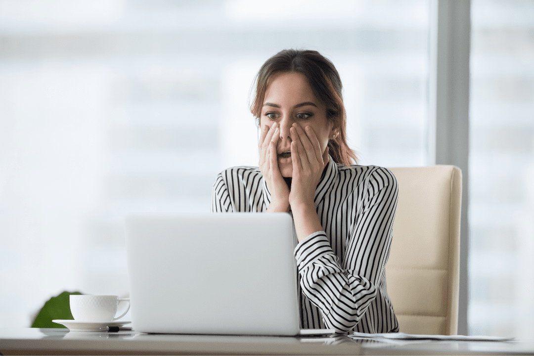 Shocked businesswoman staring at laptop with hands on face, illustrating the real-time stress and surprise of a cybersecurity breach—just like the scenario explored in the "Cyber Heist Chronicles" webinar.