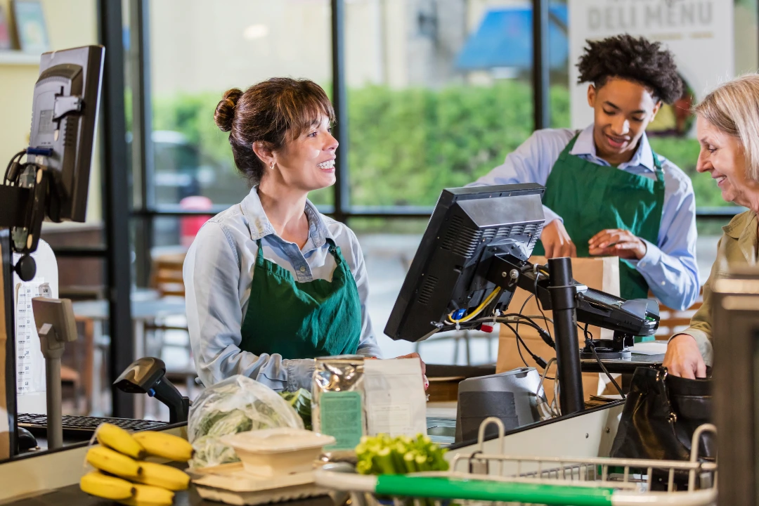 Two hourly employees in aprons helping a smiling customer, representing an example of the positive workplace culture hourly workers want to be part of.