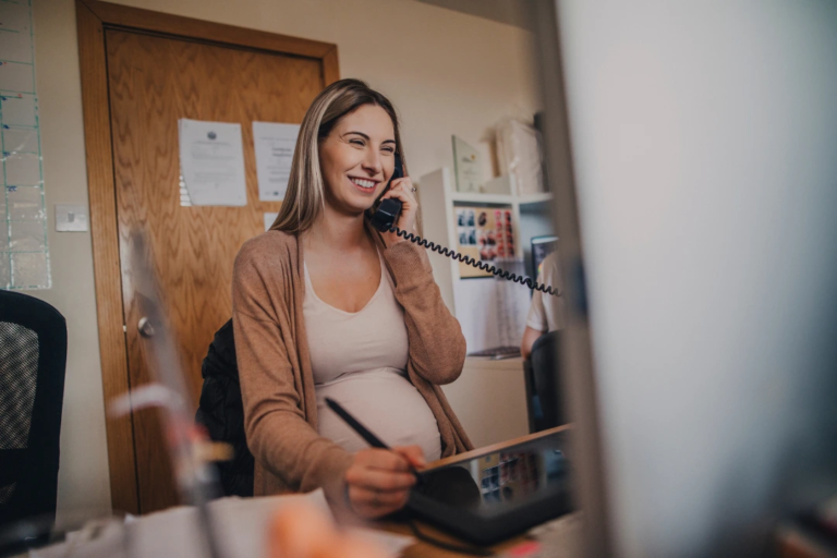 Pregnant employee smiling while speaking on the phone at her desk, representing workplace support and the importance of PWFA compliance for fair accommodations.